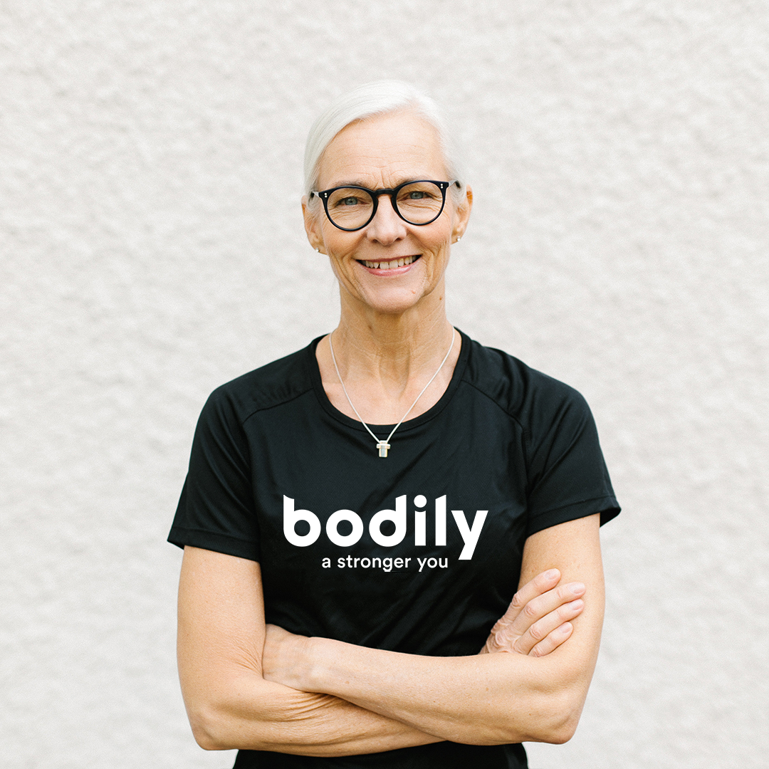 Head Coach i Bodily Karin Franzén Bohman står framför en vit vägg med armarna i kors. Har en svart t-shirt med loggan Bodily - a stronger you.
